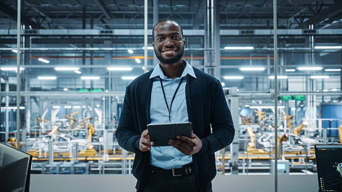 Factory Office: Portrait of Successful Black Male Chief Engineer Using Tablet Computer in Automated Robot Arm Assembly Line Manufacturing High-Tech Electric Vehicles. Medium Looking at Camera.